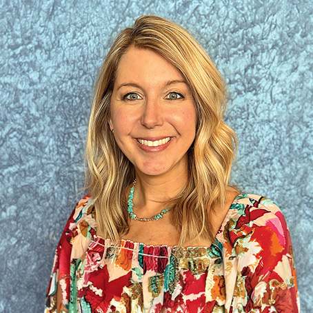 A woman with blonde hair, wearing a colorful floral top and a turquoise necklace, smiles in front of a textured blue background.