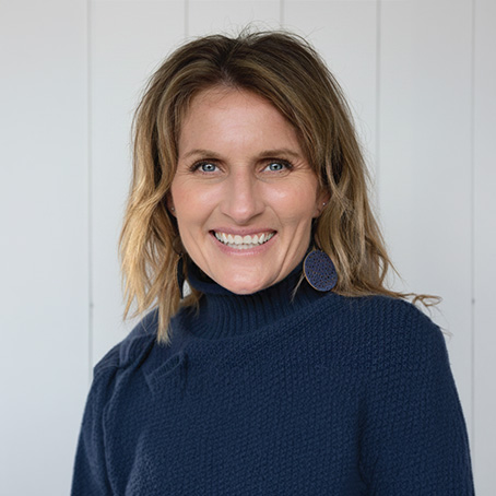 A woman with wavy light brown hair wears a dark blue textured sweater and large round earrings, smiling in front of a white shiplap wall.