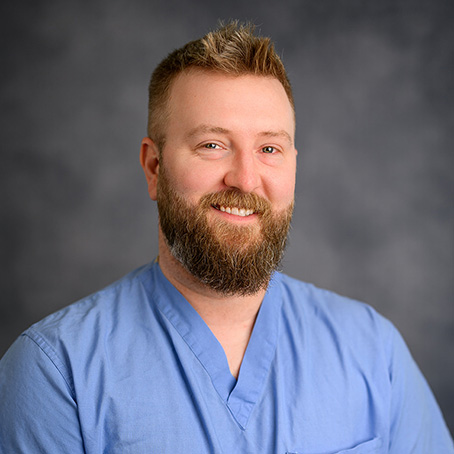 A man with short blond hair and a full beard is smiling, wearing light blue medical scrubs, standing against a plain dark gray background.