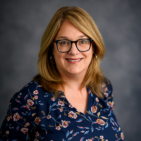 Smiling woman with shoulder-length blonde hair and glasses, wearing a navy blue floral blouse, posed in front of a gray, softly blurred background.