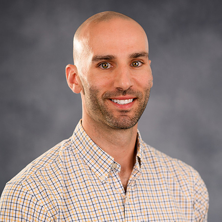 A man with a shaved head and trimmed beard, wearing a light-colored checkered shirt, smiles while posing in front of a blurred gray background.