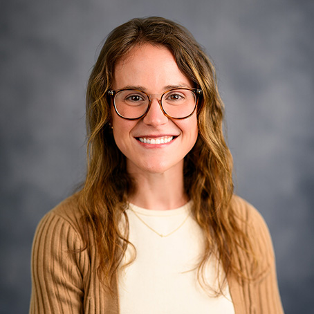 A woman with long, wavy brown hair and glasses smiles at the camera. She is wearing a beige sweater over a white top, with a simple necklace, and a gray blurred background.