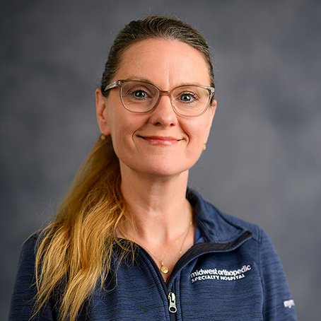 A woman with long blonde hair, glasses, and a blue zip-up top that reads “midwest orthopaedic specialty hospital,” smiling in front of a gray background.